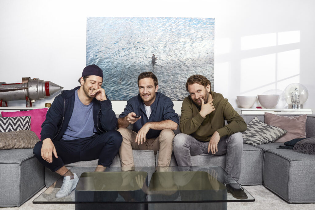 Drei junge Männer sitzen auf einer Couch und grinsen in die Kamera, die an der Position eines TV-Geräts steht. Einer der Männer bedient eine Fernbedienung.